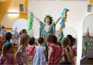 A woman dressed in a green flowing dress, holding up a garland of colourful ribbons, surrounded by children.