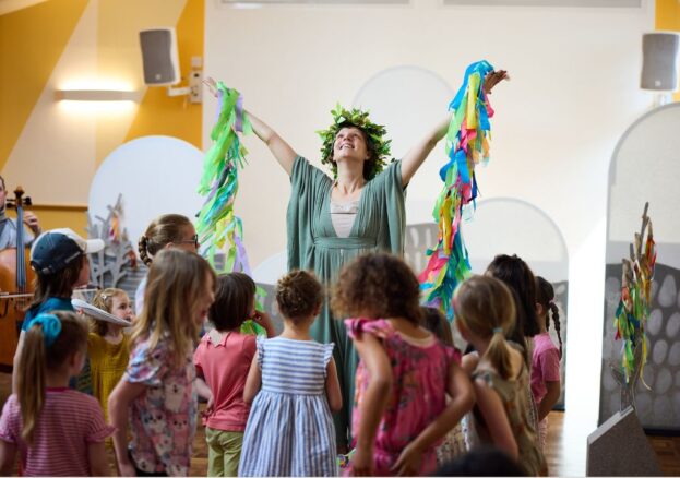 A woman dressed in a green flowing dress, holding up a garland of colourful ribbons, surrounded by children.