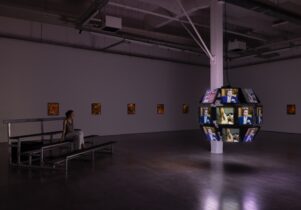 A person sits on metal bleachers looking at a hanging sphere made up of multiple television screens.
