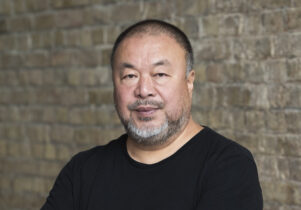Ai Weiwei lokks to camera in a black T-shirt infront of a brick wall