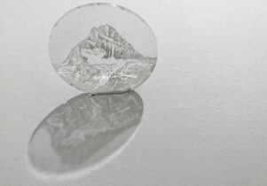 A small circular glass disc etched with a drawing of a mountain on it in black and white. It is attached to a wall and gives a shadow underneath.