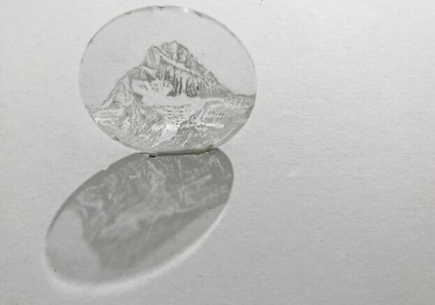 A small circular glass disc etched with a drawing of a mountain on it in black and white. It is attached to a wall and gives a shadow underneath.