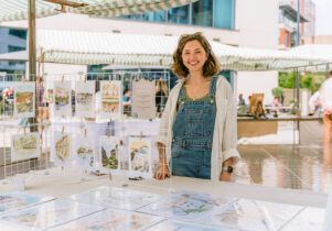 A street vendor standing behind a table selling arts and crafts.