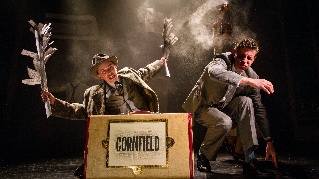 Two figures crouch on a misty stage with a suitcase that says Cornfield. One holds two paper crops and the other closes his eyes.