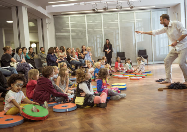 A photo taken in an event space in Central Library, in which a man performs for a group of families with young children, who are dressed in bright colours.