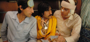 Three men sit next to each other. One's head is bandaged, one holds a torch and one wears a sleepmask.
