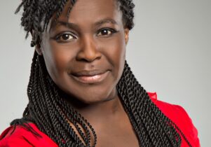 An image of Dame Maggie Aderin-Pocock, a black middle-aged woman with long hair in a red top, looking directly at the camera with a warm expression. Look for a book