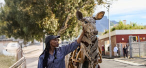 A woman in a cap touches the head of a life-sized giraffe puppet made from cardboard and wood, as part of The Herds. The background shows an urban environment with people watching and a scenic view of trees and mountains.