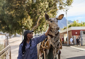 A woman in a cap touches the head of a life-sized giraffe puppet made from cardboard and wood, as part of The Herds. The background shows an urban environment with people watching and a scenic view of trees and mountains.