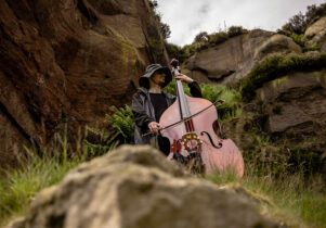 A man in a black mac and black rain hat plays a double bass on some rocks in the countryside.