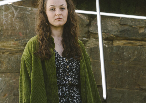 A woman wearing a long green jacket and a speckled dress stood infront of a wall.