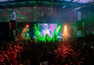 A live music performance in a dark indoor venue with colourful stage lighting and a large crowd enjoying the show. The stage features a projection screen displaying visuals, with green and orange lights illuminating the band.