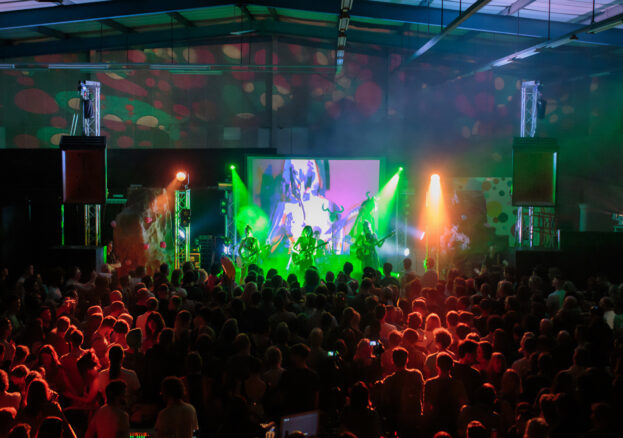 A live music performance in a dark indoor venue with colourful stage lighting and a large crowd enjoying the show. The stage features a projection screen displaying visuals, with green and orange lights illuminating the band.