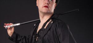 A man wearing red eye makeup dressed in black with a top hat and a cane over his shoulder.