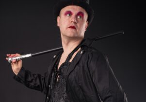 A man wearing red eye makeup dressed in black with a top hat and a cane over his shoulder.