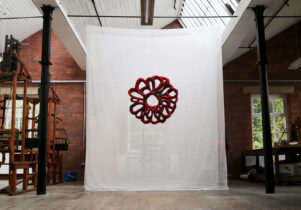 In a large industrial space filled with dobby looms hangs a blood red textile piece infront of a sheer white backdrop.