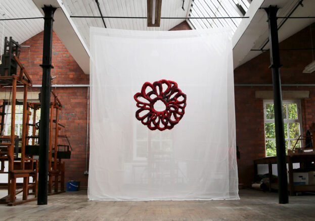 In a large industrial space filled with dobby looms hangs a blood red textile piece infront of a sheer white backdrop.