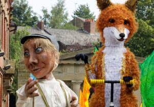 Two giant puppets: a man in overalls wearing a cap and a bright orange fox on a scooter