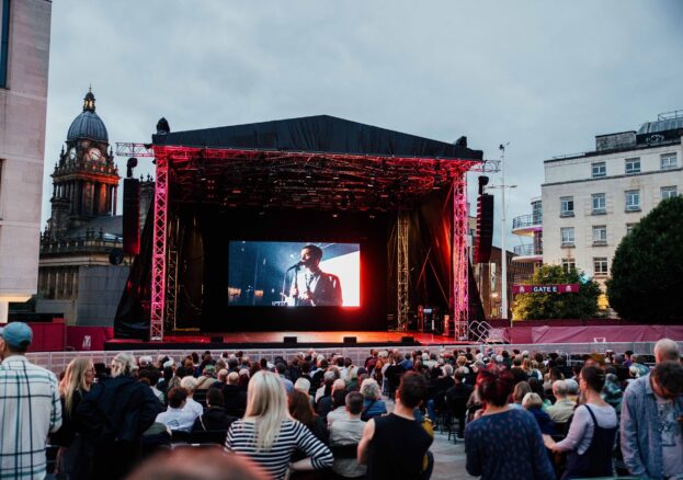 A crowd of people stand in front of a large concert tent with an illuminated screen inside it.