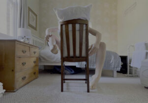 A figure in a bedroom sits backwards on a chair with a pillow covering their head and limbs contorted.