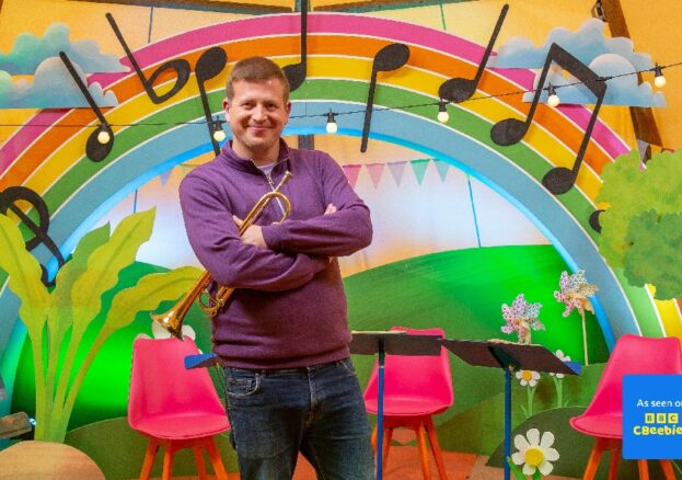 Sean Chandler stands in front of a rainbow with musical notes on it, holding a trumpet.