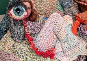 A woman sits on a crochet covered armchair, wearing a crochet costume and holding a crochet eyeball to her face