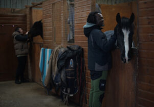 A black man and a white man are petting horses in a stable