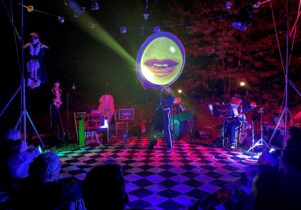 A chequered floor, a mouth projected onto a circular screen above their heads, musicians are poised to play