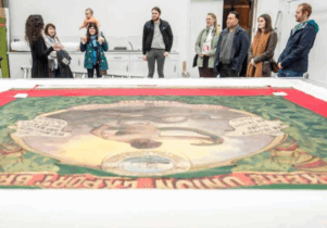 A group of people listening to a conservator, standing near a huge colourful historic banner laid out on a table.