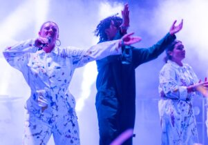 Raves R Us at Shakespeare North Playhouse: 3 performers can be seen on stage, on the left a lady wearing paint splattered overalls holds a microphone to their mouth as they sing a song. Next to them another person in blue overalls claps to the music and on the right of the image a final person wears paint splattered overalls as they also clap to the music.