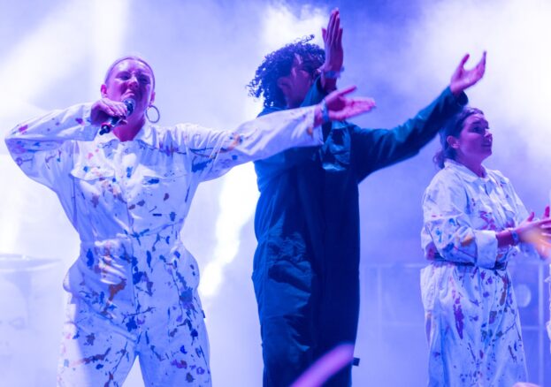 Raves R Us at Shakespeare North Playhouse: 3 performers can be seen on stage, on the left a lady wearing paint splattered overalls holds a microphone to their mouth as they sing a song. Next to them another person in blue overalls claps to the music and on the right of the image a final person wears paint splattered overalls as they also clap to the music.