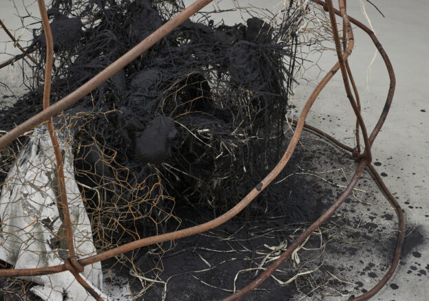 Detail of an abstract sculpture, with burned materials and rusty chicken wire at the centre, with rusted metal bars bent around it.