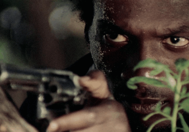 Ivan, played by Jimmy Cliff, peers through the bushes holding a handgun