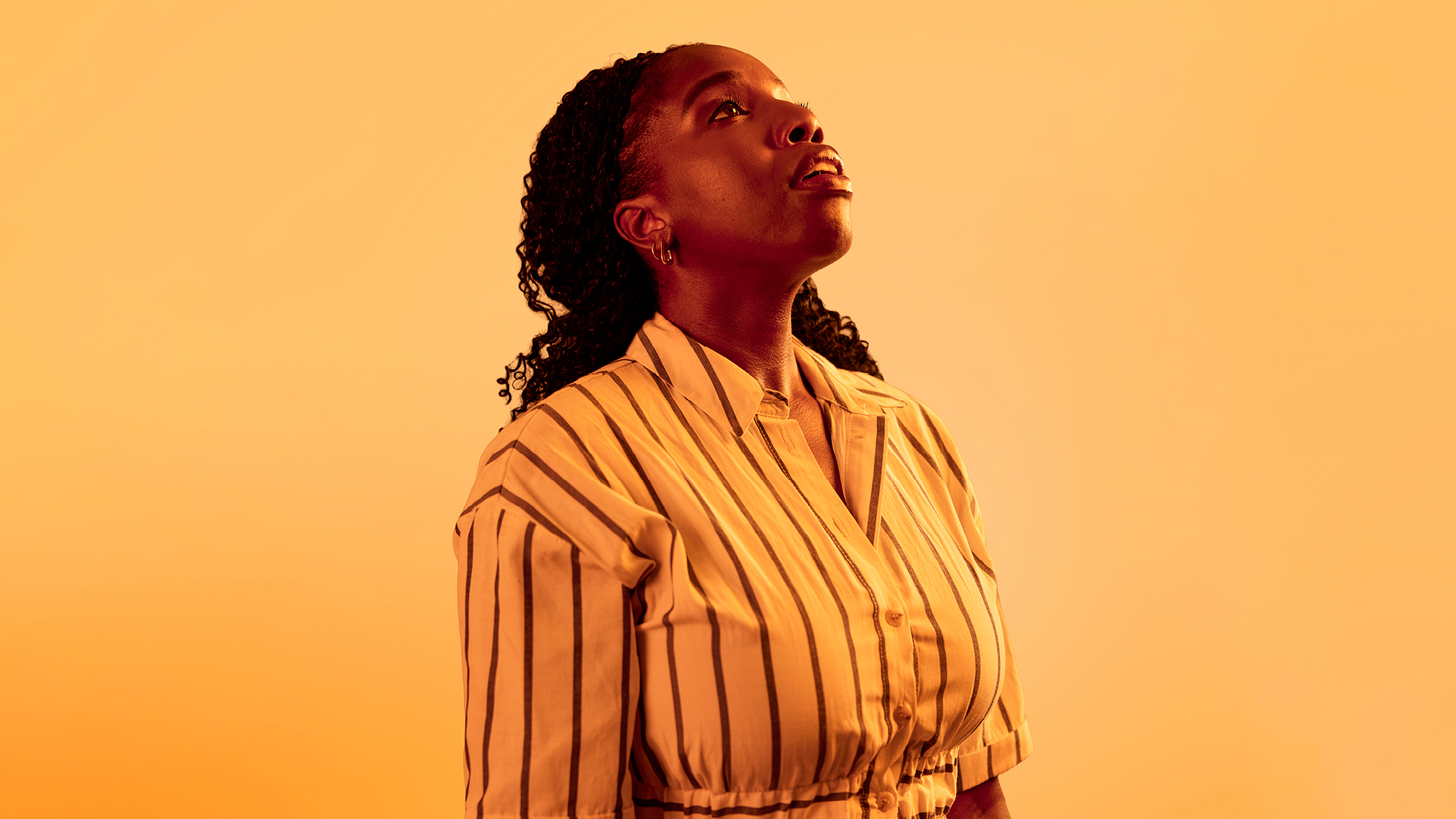 A woman of Black African heritage is drenched in yellow light.  She looks up and away from the camera as if to a sunrise. 