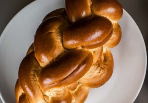  A golden-brown braided challah bread is presented on a white plate, viewed from slightly above. 