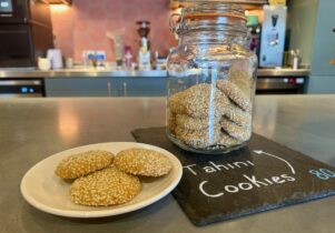A still life image displays a white plate with three round, golden-brown cookies heavily coated in sesame seeds, positioned next to a large glass jar filled with more of the same cookies. The jar sits on a dark slate board with 