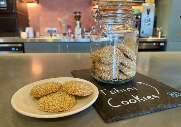A still life image displays a white plate with three round, golden-brown cookies heavily coated in sesame seeds, positioned next to a large glass jar filled with more of the same cookies. The jar sits on a dark slate board with