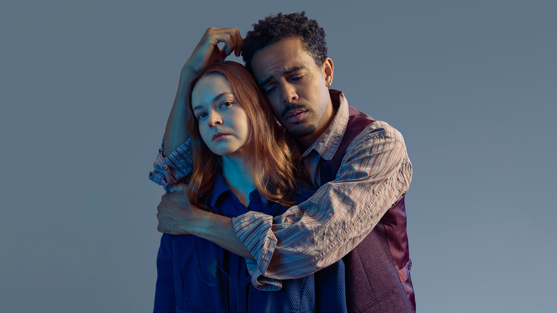 A man and woman drenched in blue light embrace. The woman looks to the camera, emotionless. The man has his eyes closed. 