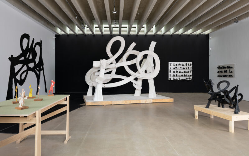 An art gallery featuring a large white abstract sculpture with looping forms at its centre. Surrounding it are smaller sculptures in various colours and styles displayed on wooden tables, against a contrasting black wall.