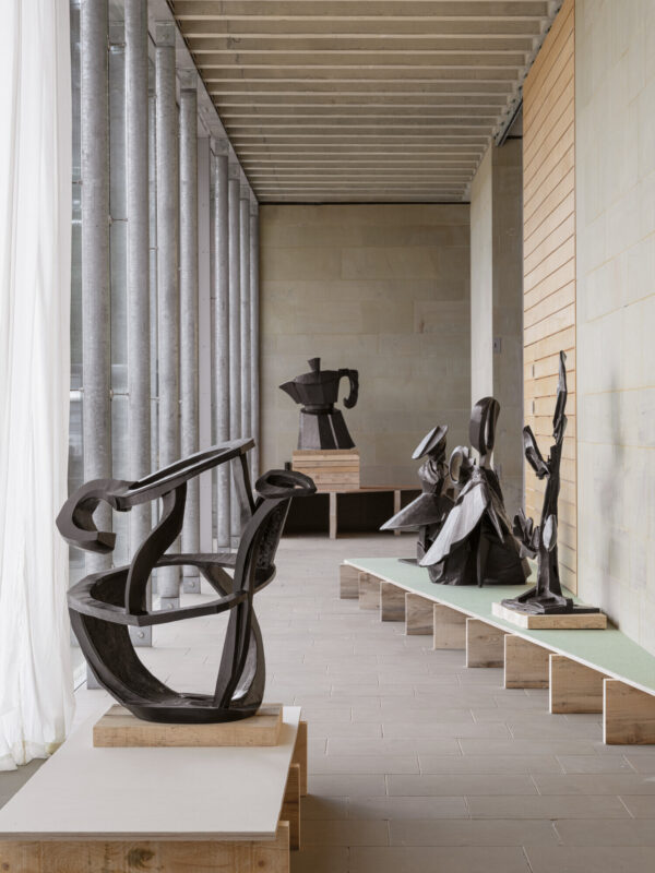 A spacious gallery features several abstract black sculptures displayed on wooden platforms. Natural light filters through large windows, illuminating the minimalist interior with stone walls and a green bench, creating a serene artistic atmosphere.