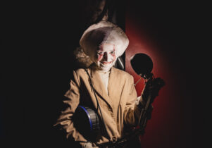 Image of a performer dressed as a creepy moon, wearing a beige suit jacket, grinning in front of a lamp