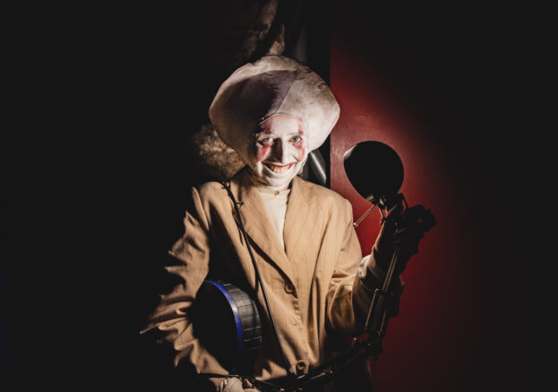 Image of a performer dressed as a creepy moon, wearing a beige suit jacket, grinning in front of a lamp