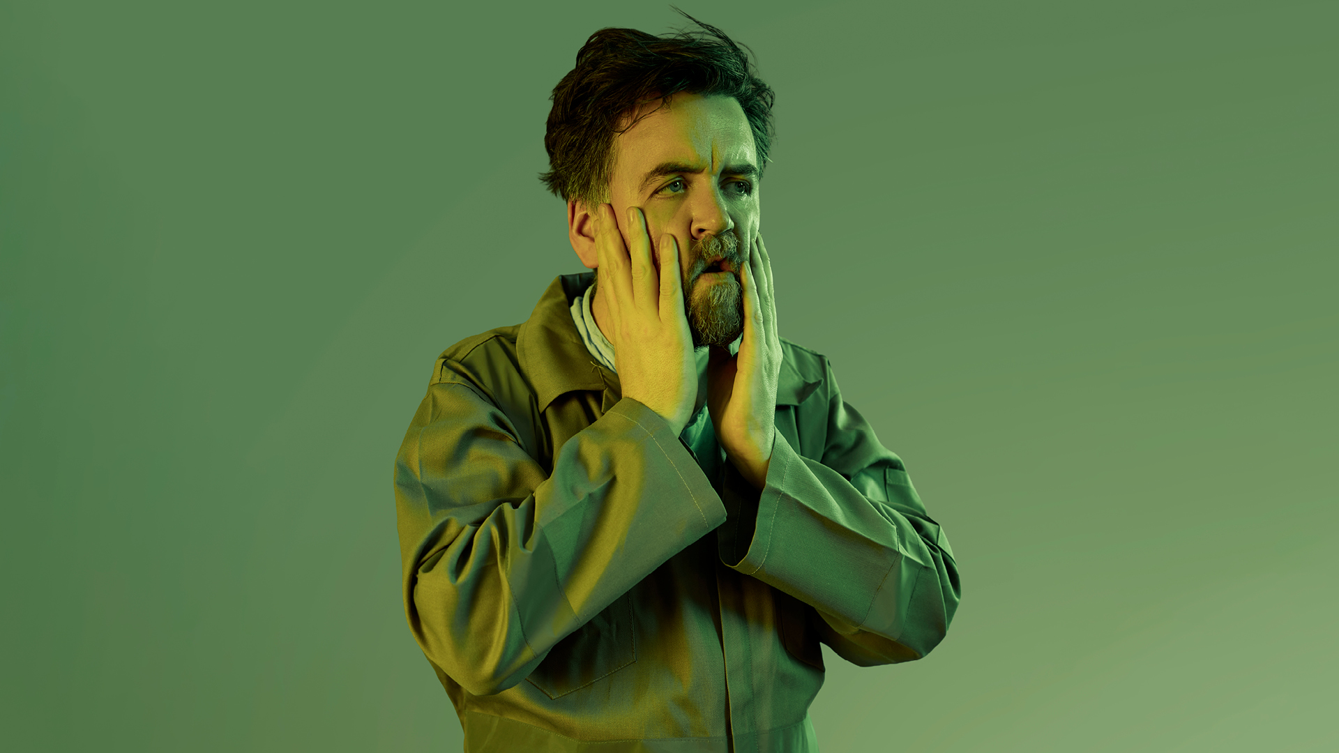 A man, drenched in green light has his head in his hands and looks beyond the camera. 