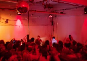 A group dances in a dimly lit room, bathed in red and orange light. A DJ, centered in the background with headphones on, plays records. The image is intentionally blurred to obscure faces.