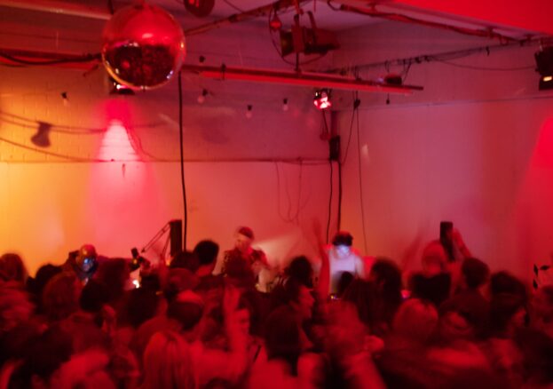 A group dances in a dimly lit room, bathed in red and orange light. A DJ, centered in the background with headphones on, plays records. The image is intentionally blurred to obscure faces.