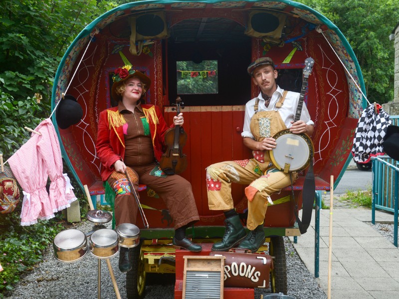 Two people with string instruments sit at the back of a red cart.