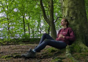 A lady sat at the base of a tree in a woodland holding a mug and looking reflectively