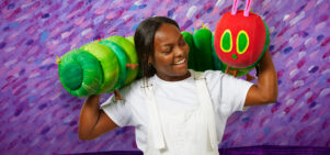 A person stands with The Very Hungry Caterpillar puppet resting across their shoulders, the background is purple.
