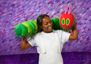A person stands with The Very Hungry Caterpillar puppet resting across their shoulders, the background is purple.