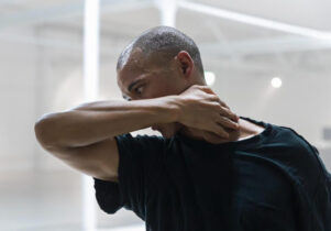 blackmilk at Stanley & Audrey Burton Theatre: A black man is pictured in motion with his profile central to the image, he is visible only from the waist up. The room is brightly lit and has white industrial features. He has short, dark, afro hair and is wearing an oversized black t-shirt. His right arm extends out in front of him, bending at the elbow, his hand reaches around, resting on the nape of his neck.